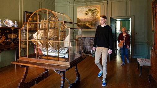A large model ship with intricate sails and guns detail inside a glass cabinet on display at Treasurers House. Two visitors are walking into the dark wood floor and green walled room to look at it.